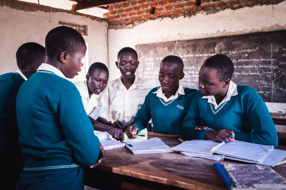 Six youth look at and discuss at notes inside a school. © Giovanni Okot