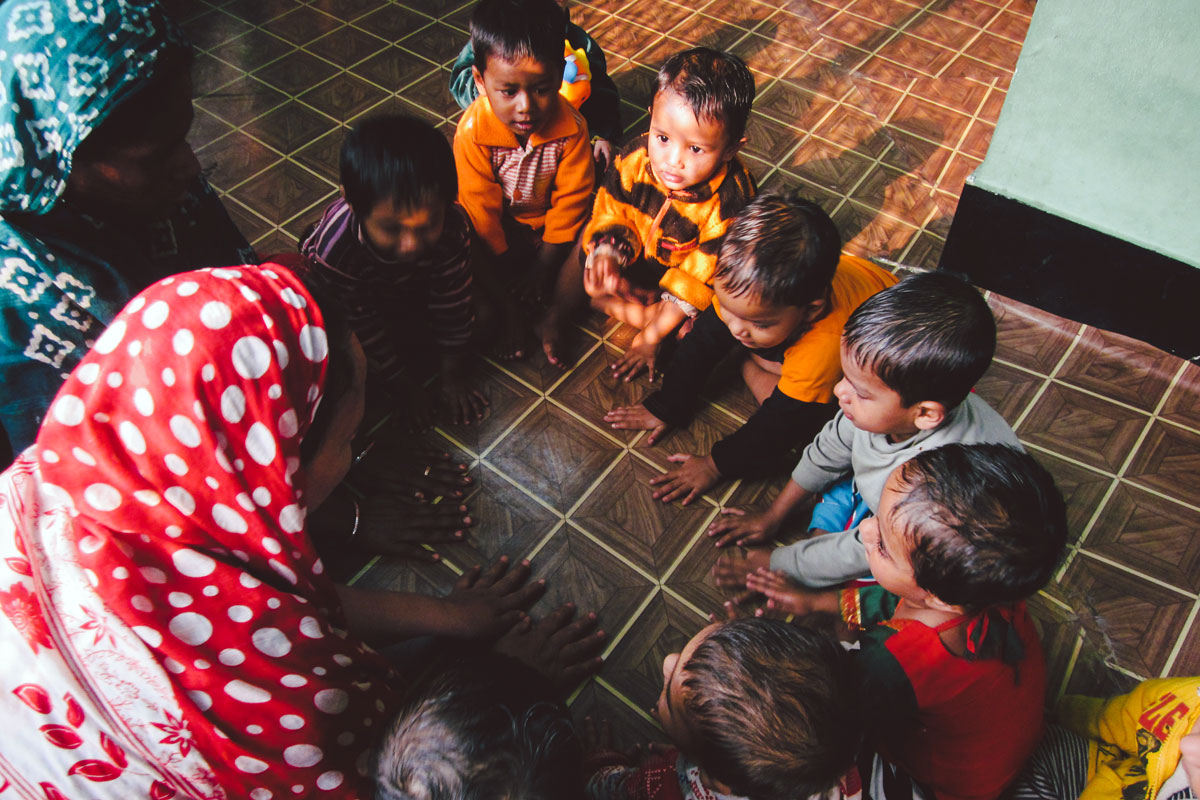 Young children sit in a circle on the floor playing a game with two teachers.