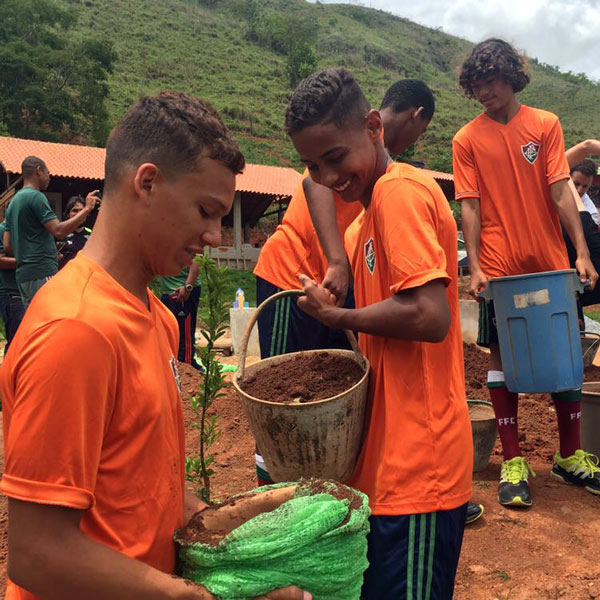 Four youth in orange shirts hold buckets with soil and plants outside. One smiles broadly while looking at the bucket.