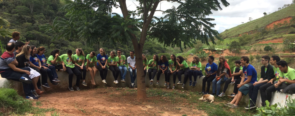 About 30 young people in green and blue shirts sit and talk on a concrete, semicircular bench outside, underneath a tree.