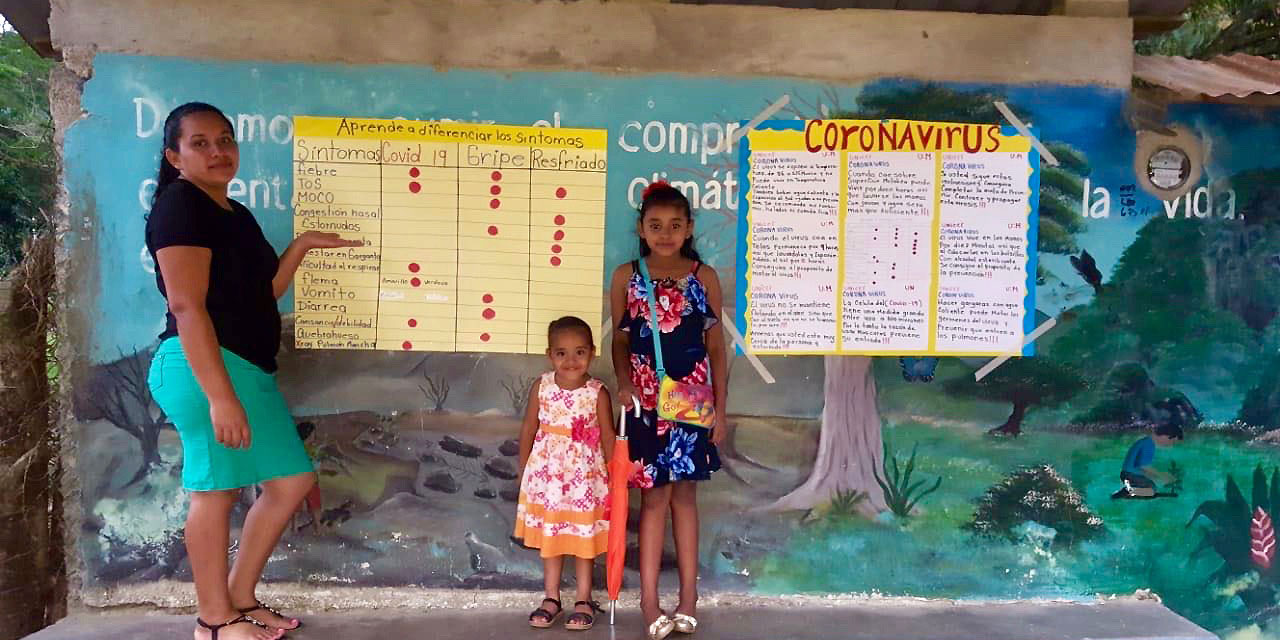 A woman and two children stand in front of posters with information about coronavirus.