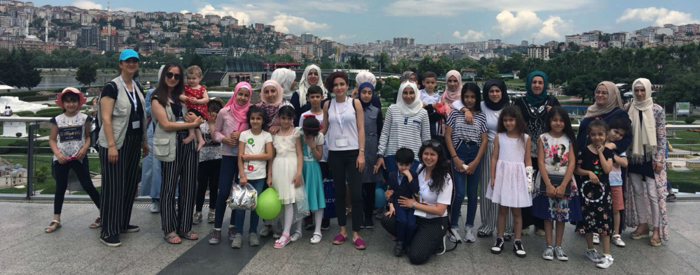 A group of women and children pose for a photo on a wide, tiled walkway, with a city and cloudy sky behind them.