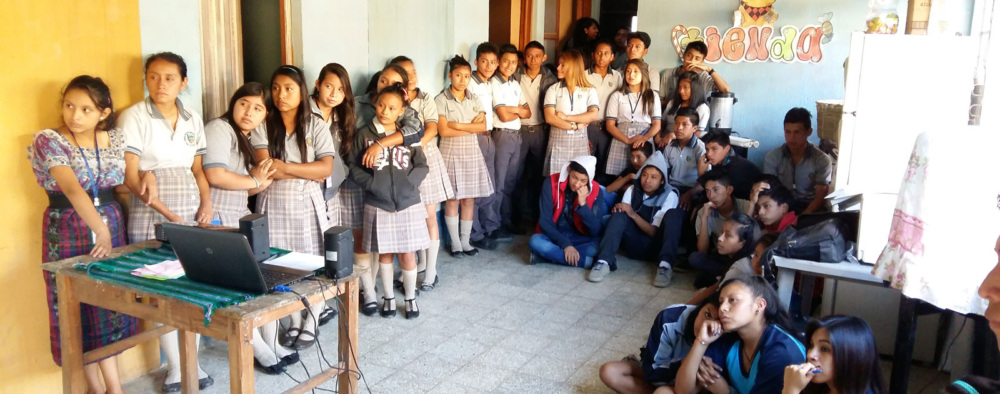 A group of adolescents sit and stand in a circle in a classroom, watching a presentation.