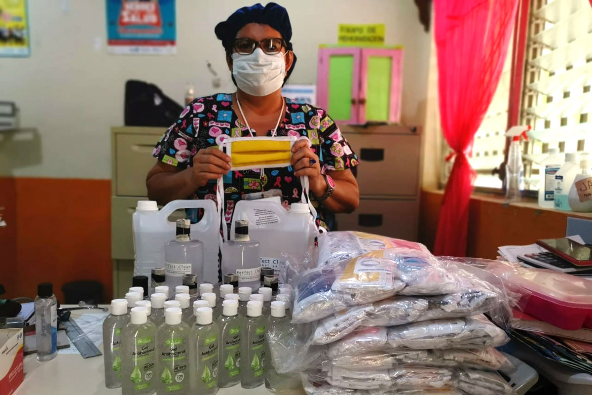 A woman wearing a medical face mask stands behind hand sanitizer and emergency supplies.
