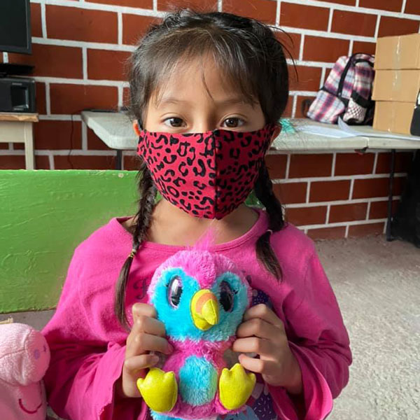 A girl wearing a mask holds a colorful stuffed animal.