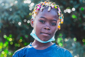 Outside, a girl with a blue shirt, colorful braids, and a lowered face mask looks at the camera.