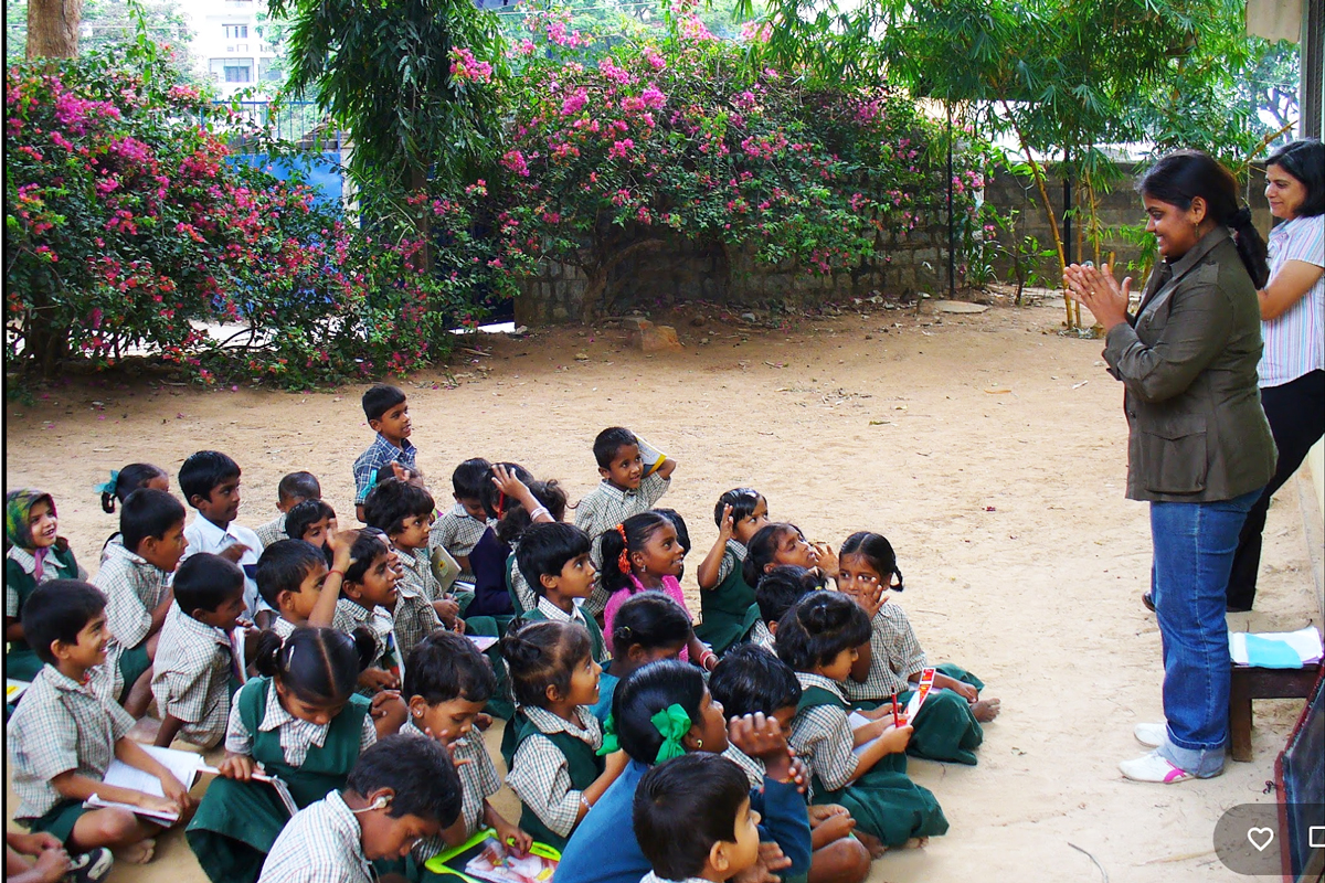A lesson being conducted for young students seated on the ground outside. 