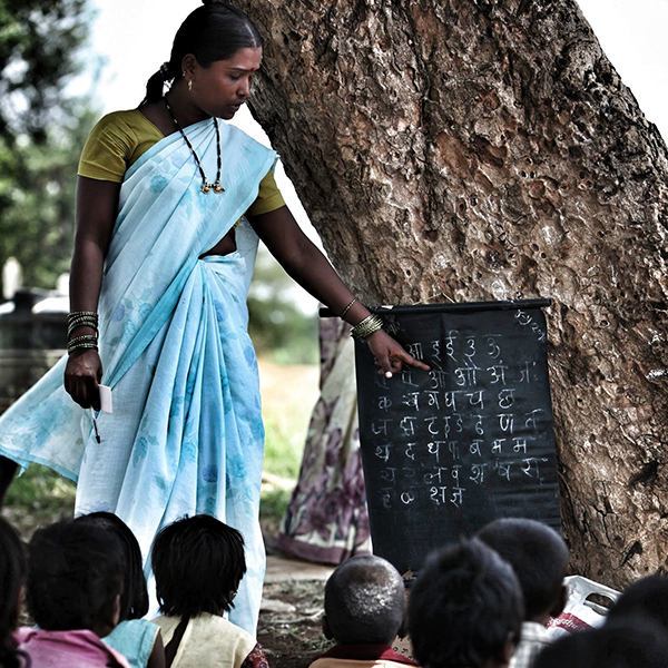 A teacher explaining something with a blackboard and chalk to young students seated outdoors