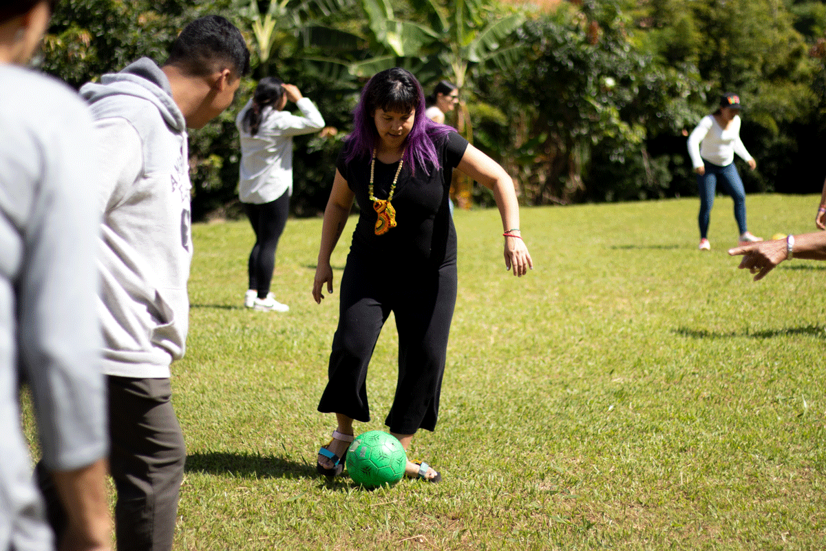 A woman dressed in black with a large necklace kicking a green football on a grassy playing field.