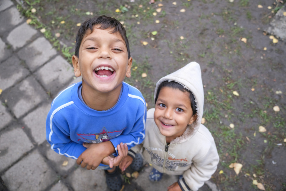 Two smiling children look up at the camera. One wears a blue sweater, and the other wears a light-colored hoodie. They stand on a stone path with grass and scattered leaves in the background.