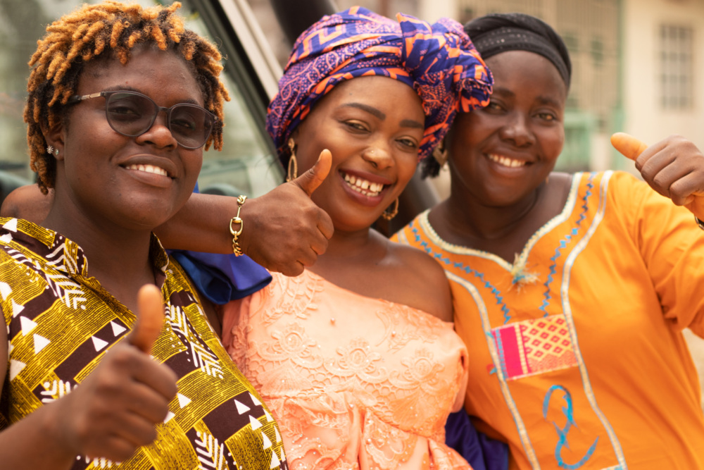 Three smiling women in colorful traditional clothing give thumbs up. One wears glasses and a patterned shirt, another is in an orange off-shoulder dress with a headwrap, and the third wears an orange dress. They stand together outdoors, radiating joy.