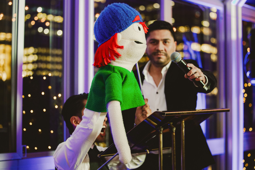 A person holds up a mic to the face of a puppet, dressed in a green tshirt and wearing a baseball cap, during a puppet show at a GFC event. 