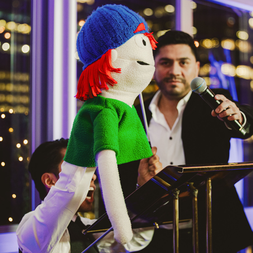 A person holds up a mic to the face of a puppet, dressed in a green tshirt and wearing a baseball cap, during a puppet show at a GFC event.