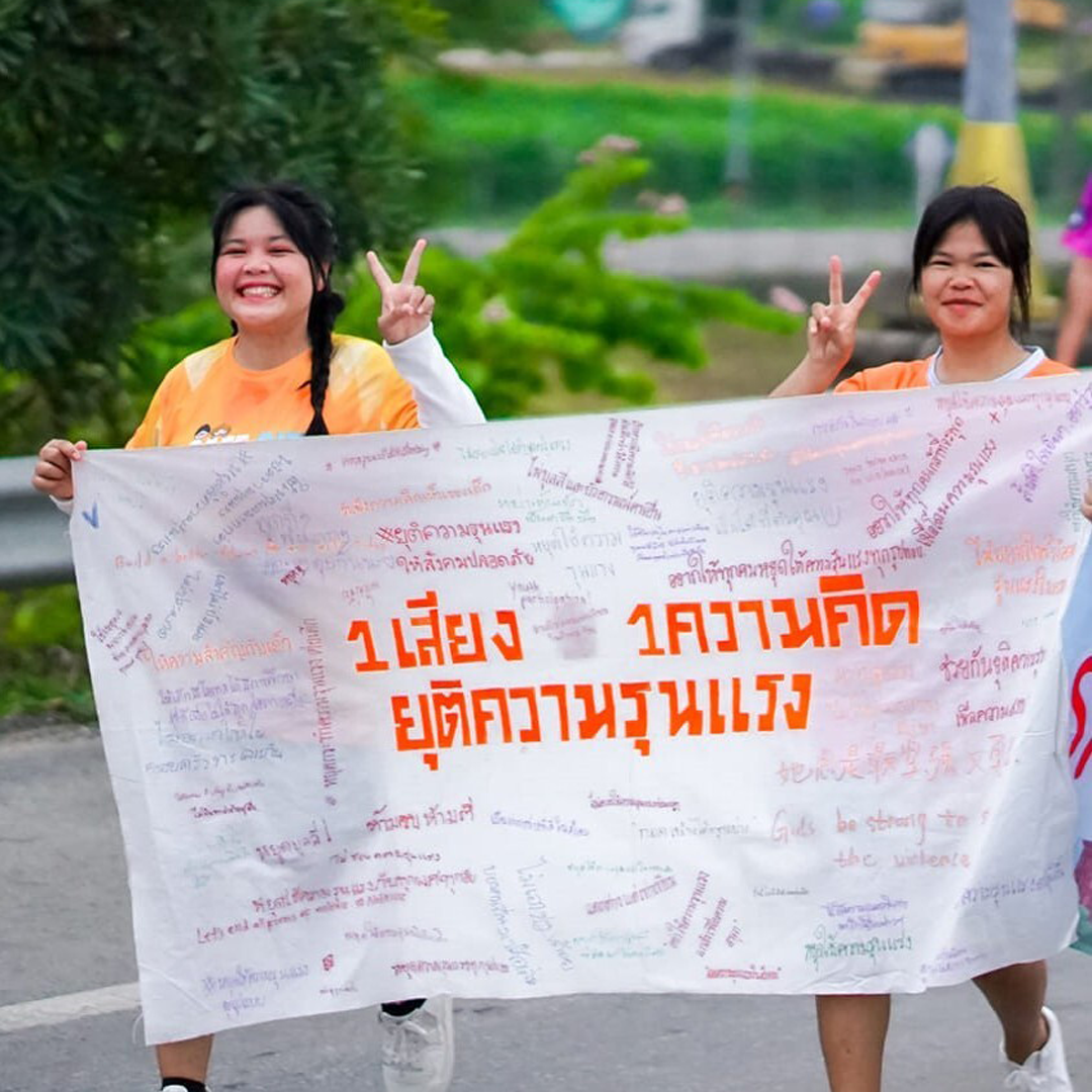 Two girls from Thailand holding a very large fan, smiling at the camera with their hands making a peace sign