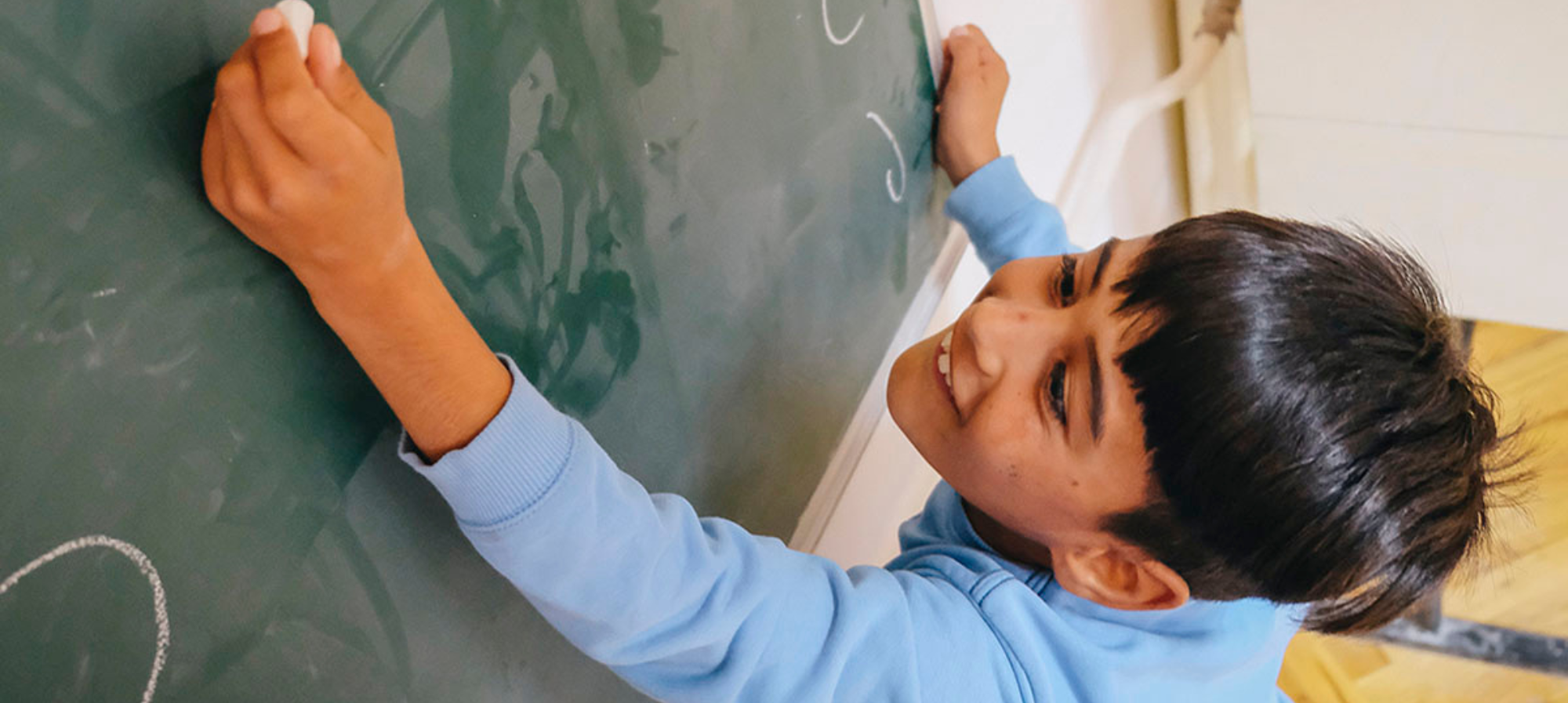 A young boy reaching up to write on the blackboard with chalk